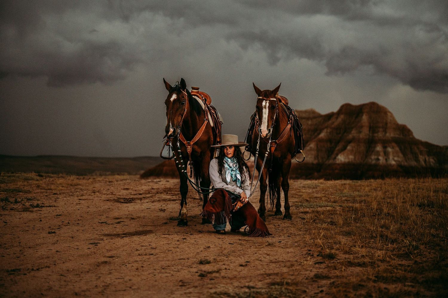 Two brown horses with riding gear stand on dusty ground against dramatic stormy sky and rocky formations.