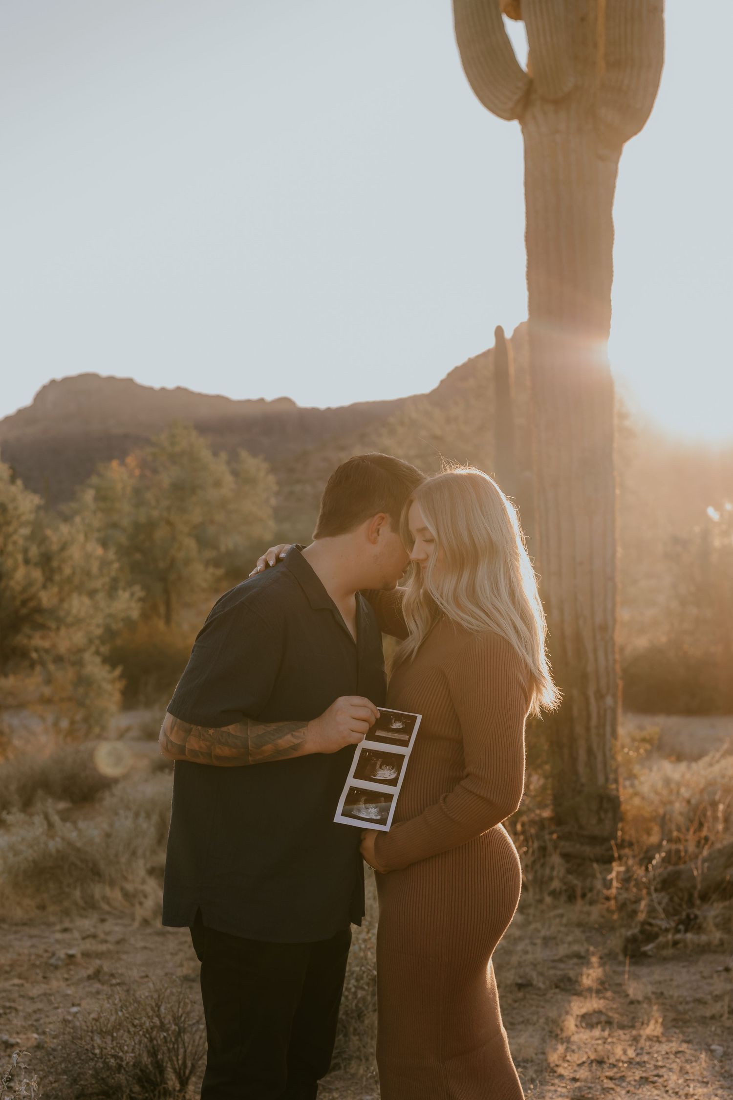 A couple embraces in front of a tall saguaro cactus at sunset while holding an ultrasound photo in the desert.