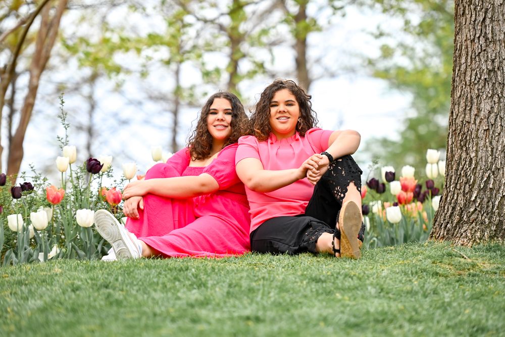 Two people in bright pink outfits sit together on grass surrounded by colorful tulips in a spring garden.
