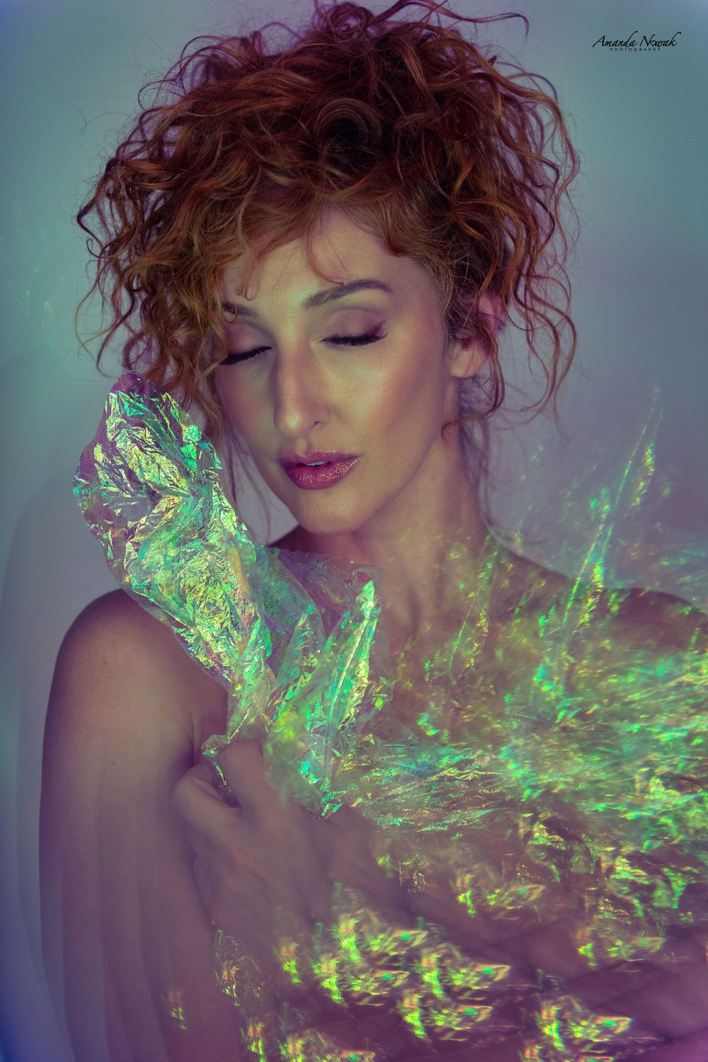 Red-haired woman with eyes closed holding iridescent holographic material in soft light.