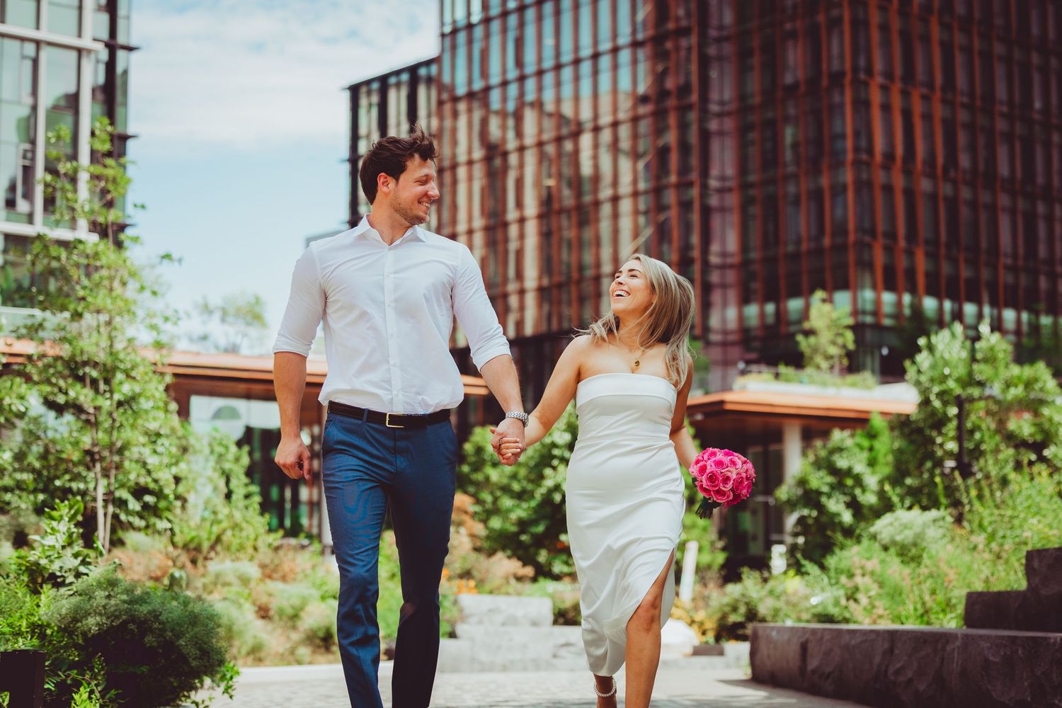 A couple holding hands while running through an urban garden path with modern buildings in the background.