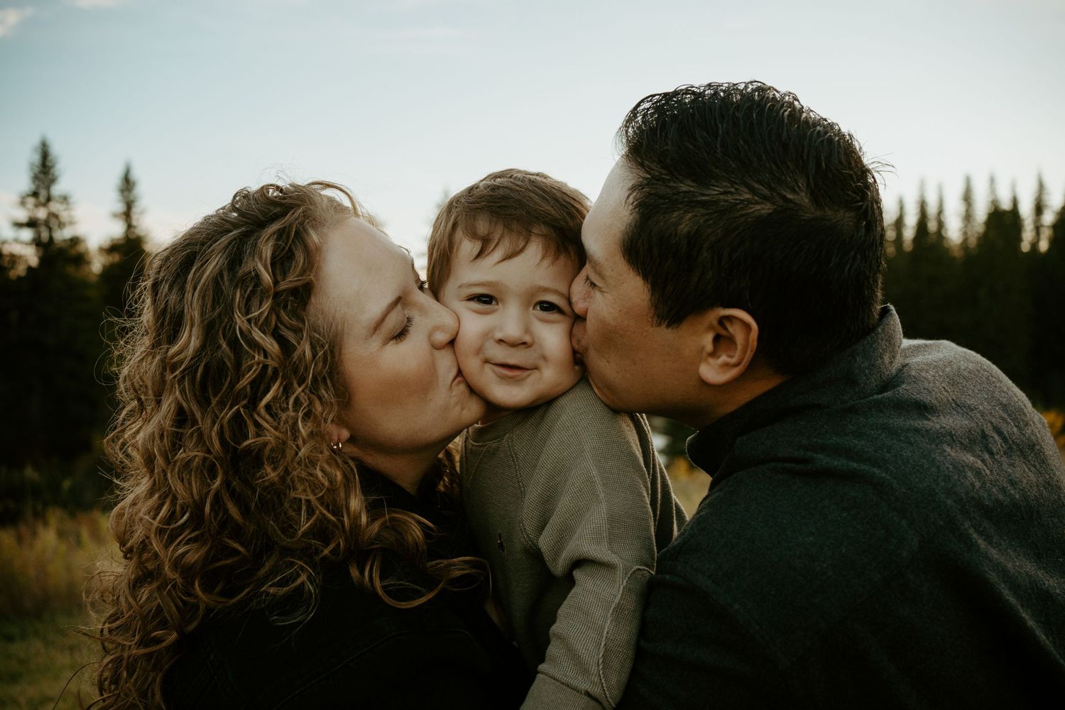 A tender family moment captured outdoors with autumn trees in the background at sunset.