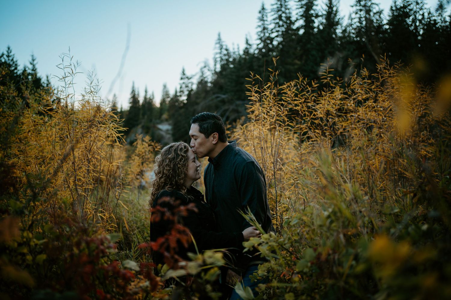 A couple shares an intimate moment surrounded by golden autumn foliage and evergreen trees in a moody forest setting.