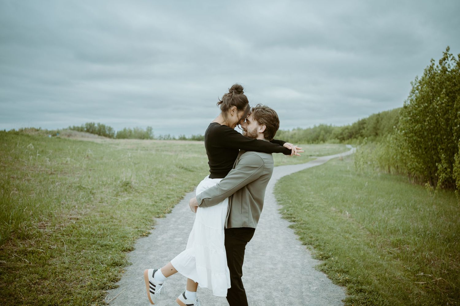A romantic embrace on a winding path through green fields under an overcast sky.