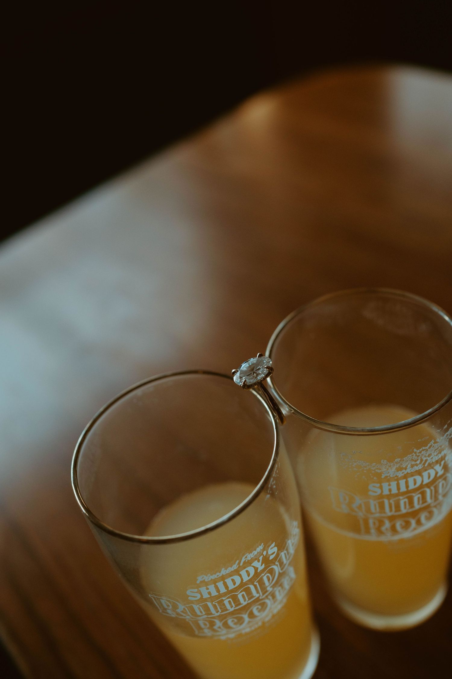 Two Shiddy's beer glasses with golden ale and an engagement ring resting on the rims on a wooden table.
