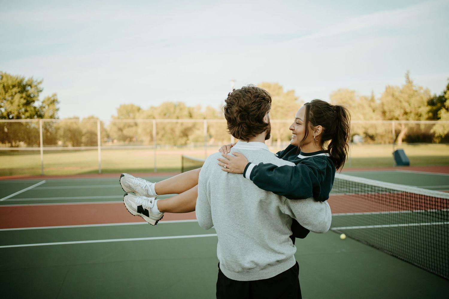 A couple embrace playfully on a tennis court during an afternoon tennis session.