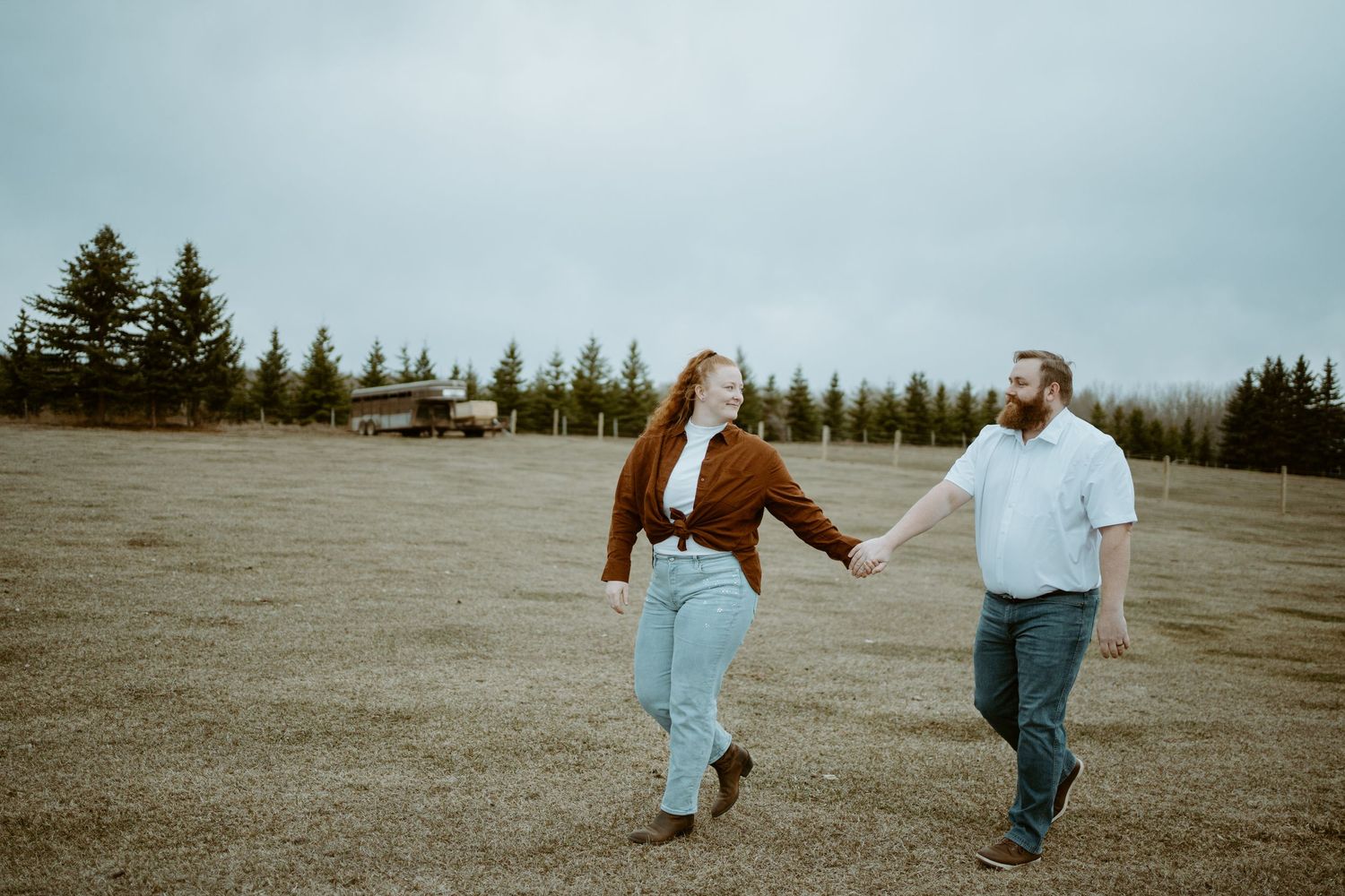 Couple walking hand in hand through an open field with evergreen trees in the background on an overcast day.