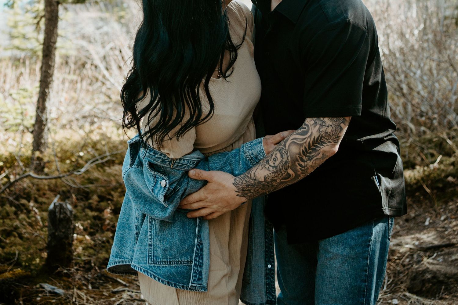 A couple embraces while wearing denim and black clothing outdoors with visible tattoo art in natural lighting.