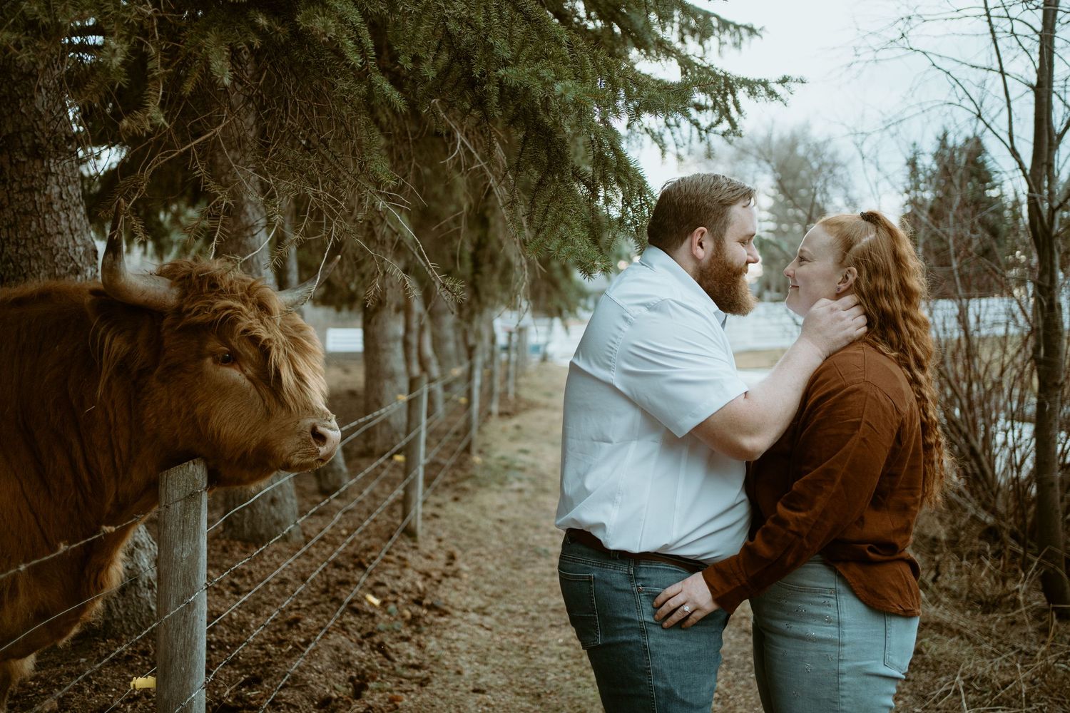 A couple dressed in casual attire stand together on a path with a curious brown horse peeking from behind a fence.