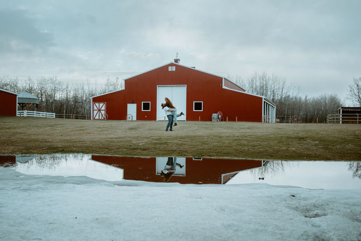 A red barn reflects perfectly in a puddle of water on a winter day, creating a mirror image against the rural landscape.