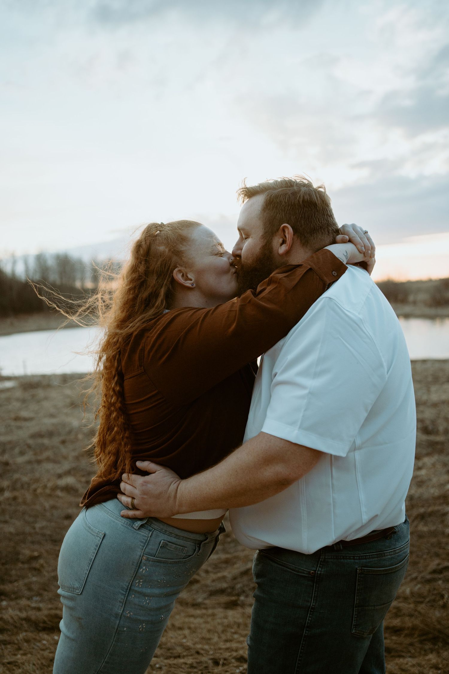 A tender romantic moment shared between a couple embracing outdoors at sunset near a lake.