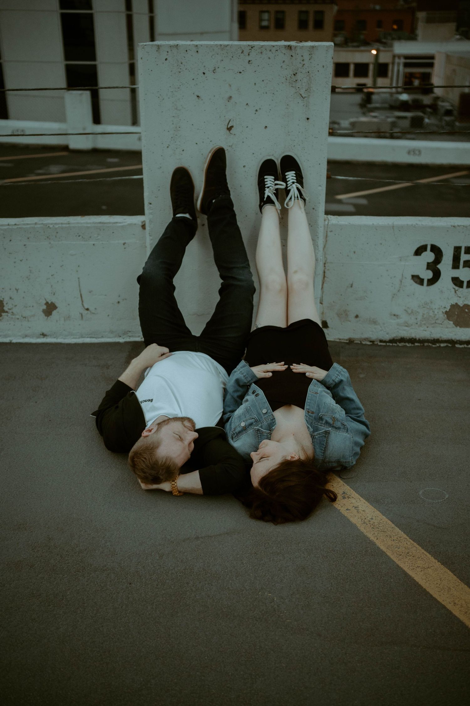 Two people lying on concrete ground with legs up against a wall wearing casual clothing and shoes in a parking lot.