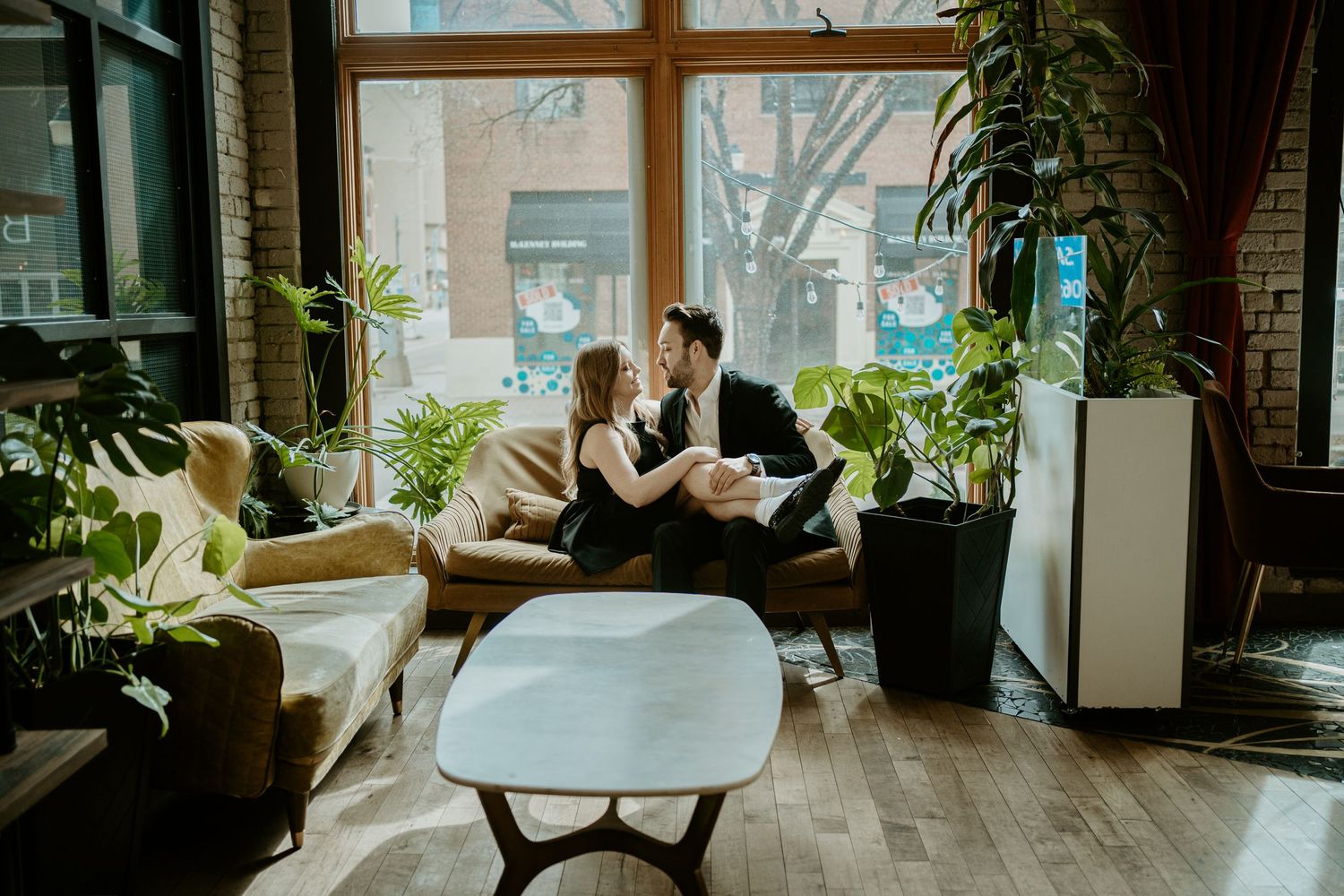 Two people sit close together on a sofa in a sunlit living room with houseplants and modern decor.