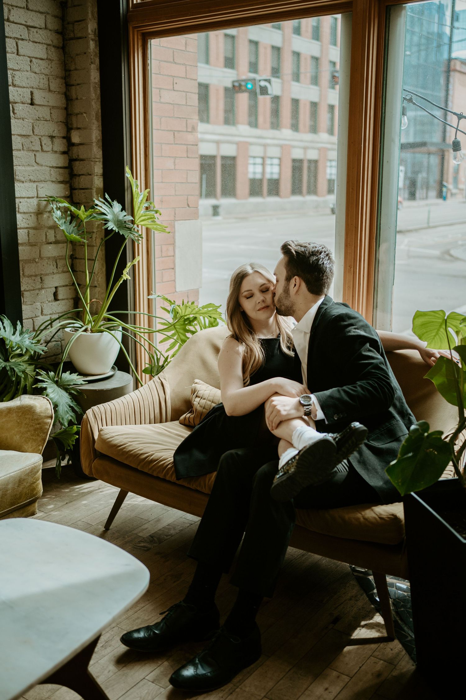 A romantic couple shares an intimate moment on a vintage couch by a window surrounded by houseplants in an urban setting.