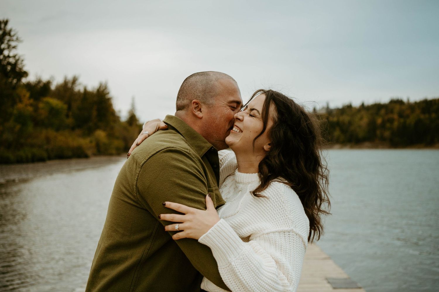 A couple in wedding attire shares romantic kisses by a serene lakeside on an autumn afternoon.