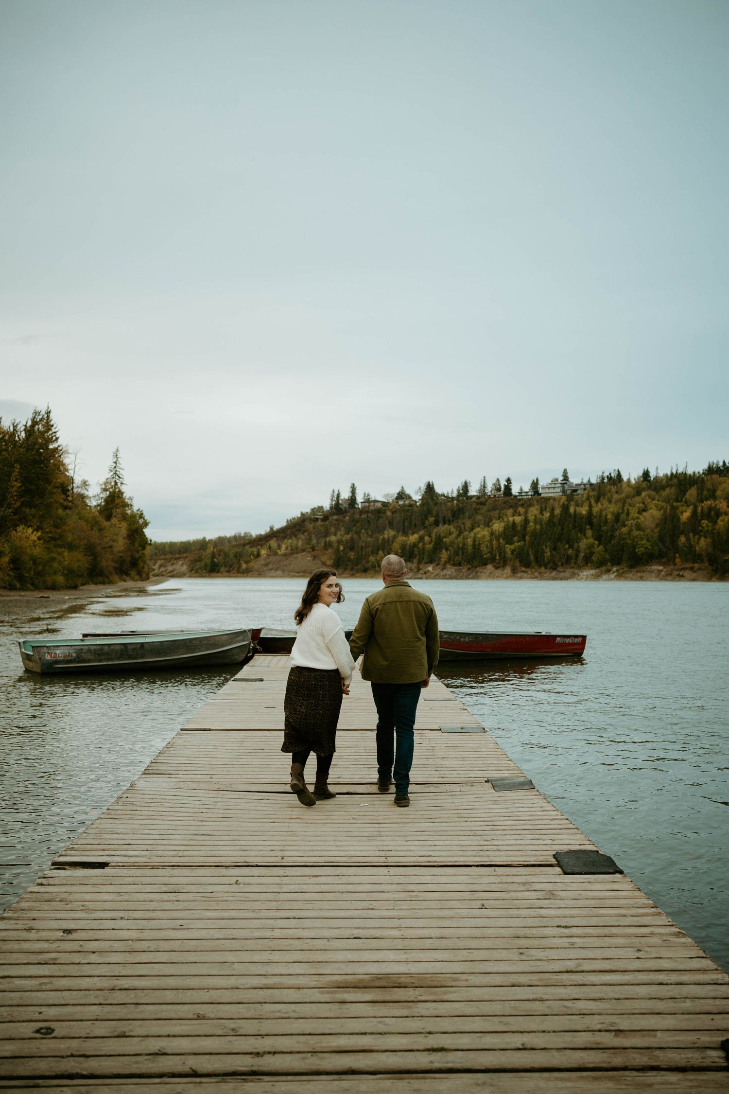 A couple walks together on a wooden dock overlooking a serene lake with boats and evergreen trees in the background.