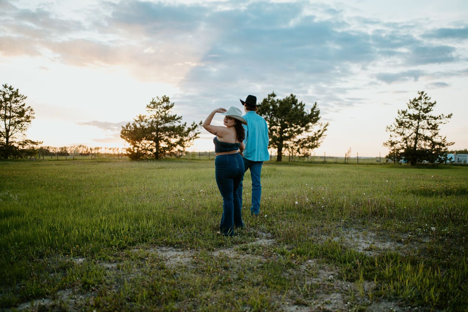 A couple stands together in an open field at sunset with silhouetted pine trees in the background.
