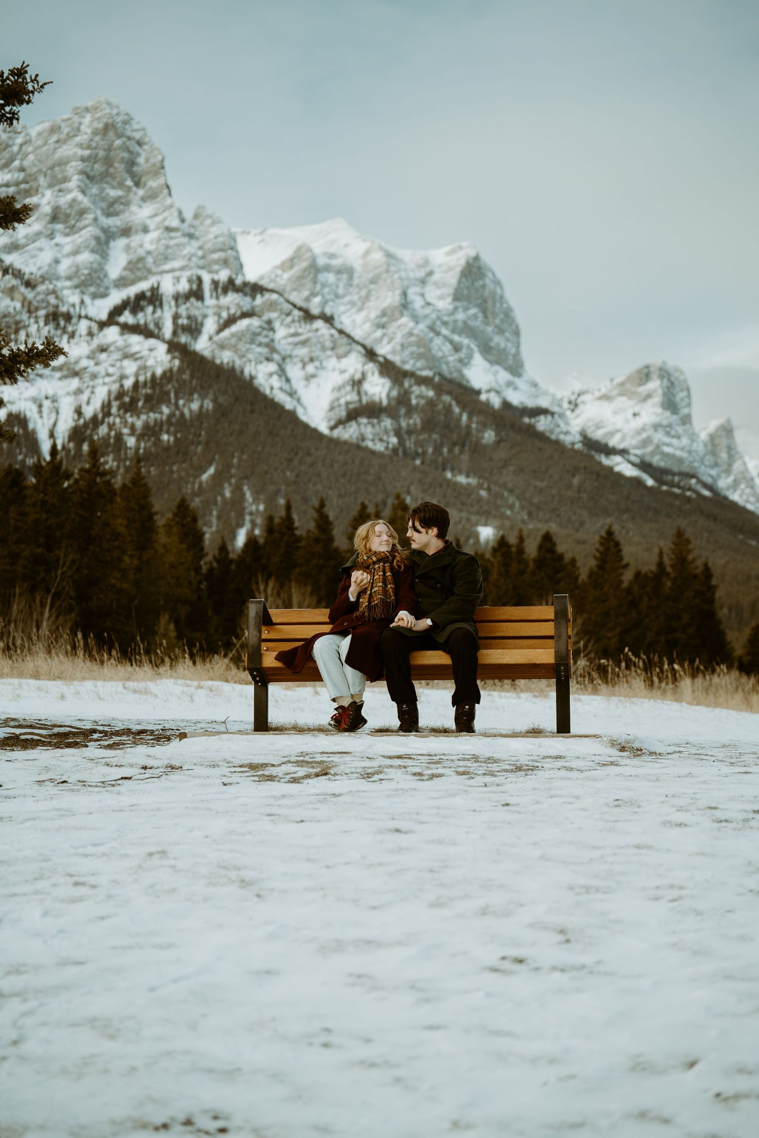A couple sits on a wooden bench in a snowy landscape with majestic snow-capped mountain peaks rising in the background.
