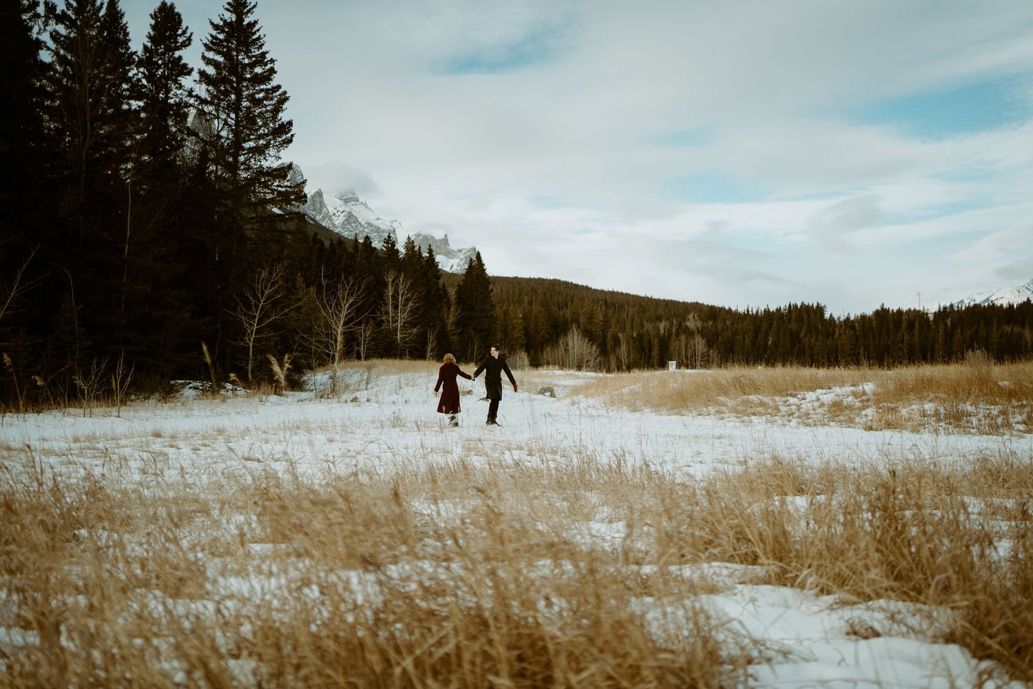 Two figures walk together across a snow-covered winter meadow surrounded by evergreen forest on a cloudy afternoon.