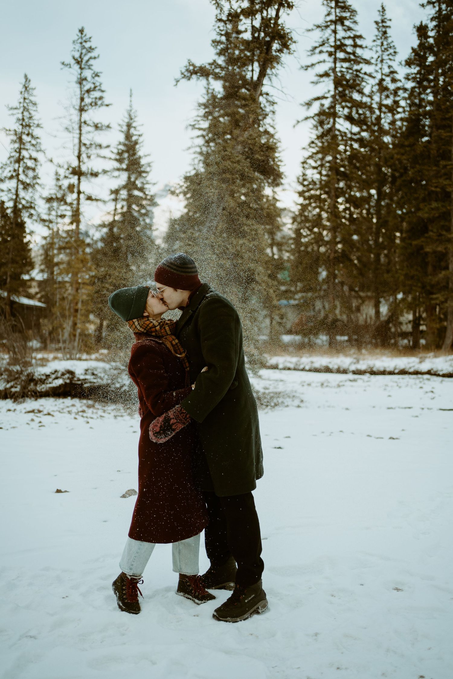 A couple embraces in winter coats while standing in snowy landscape surrounded by evergreen trees at dusk.