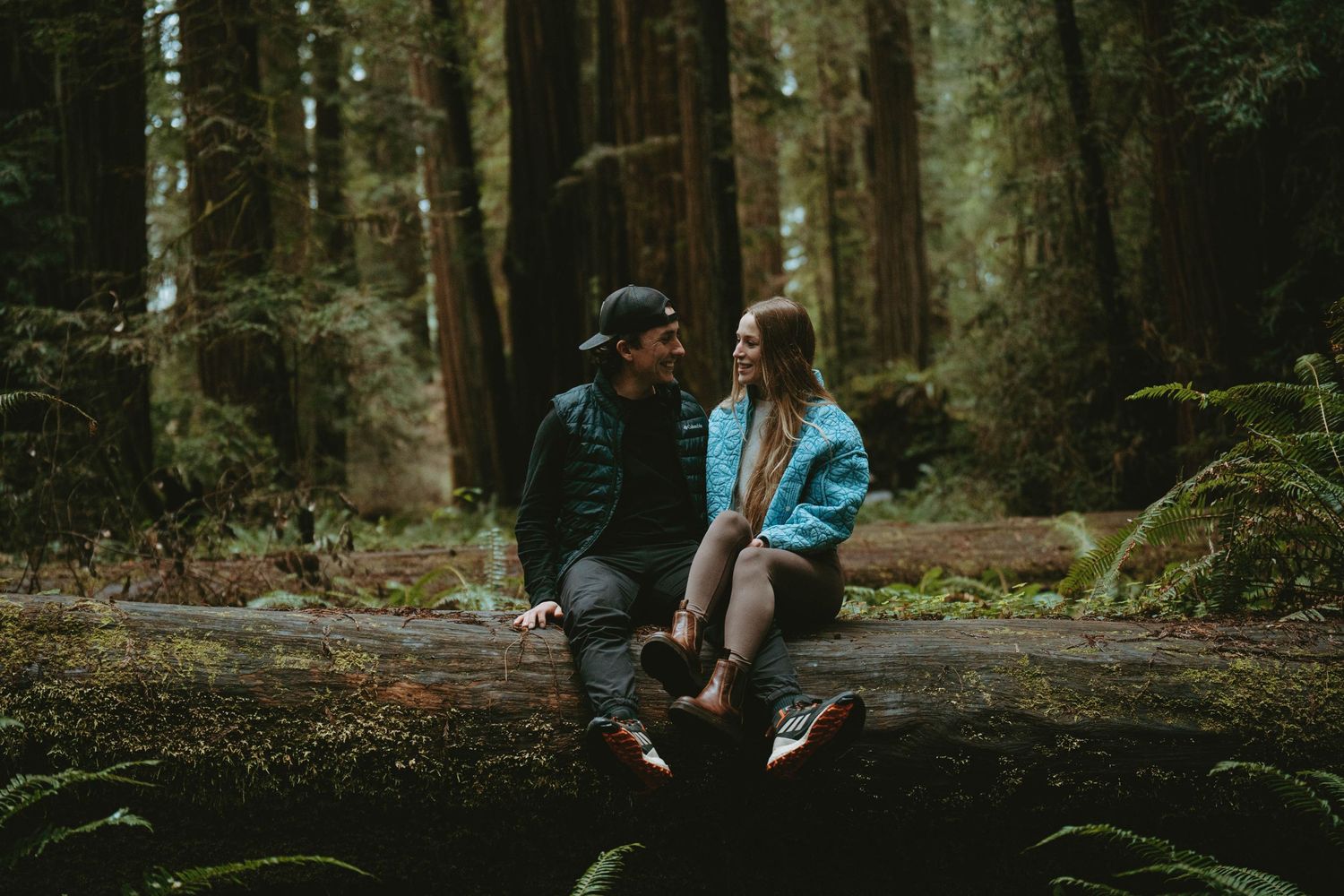 Two people sitting together on fallen log in misty forest wearing outdoor clothing.