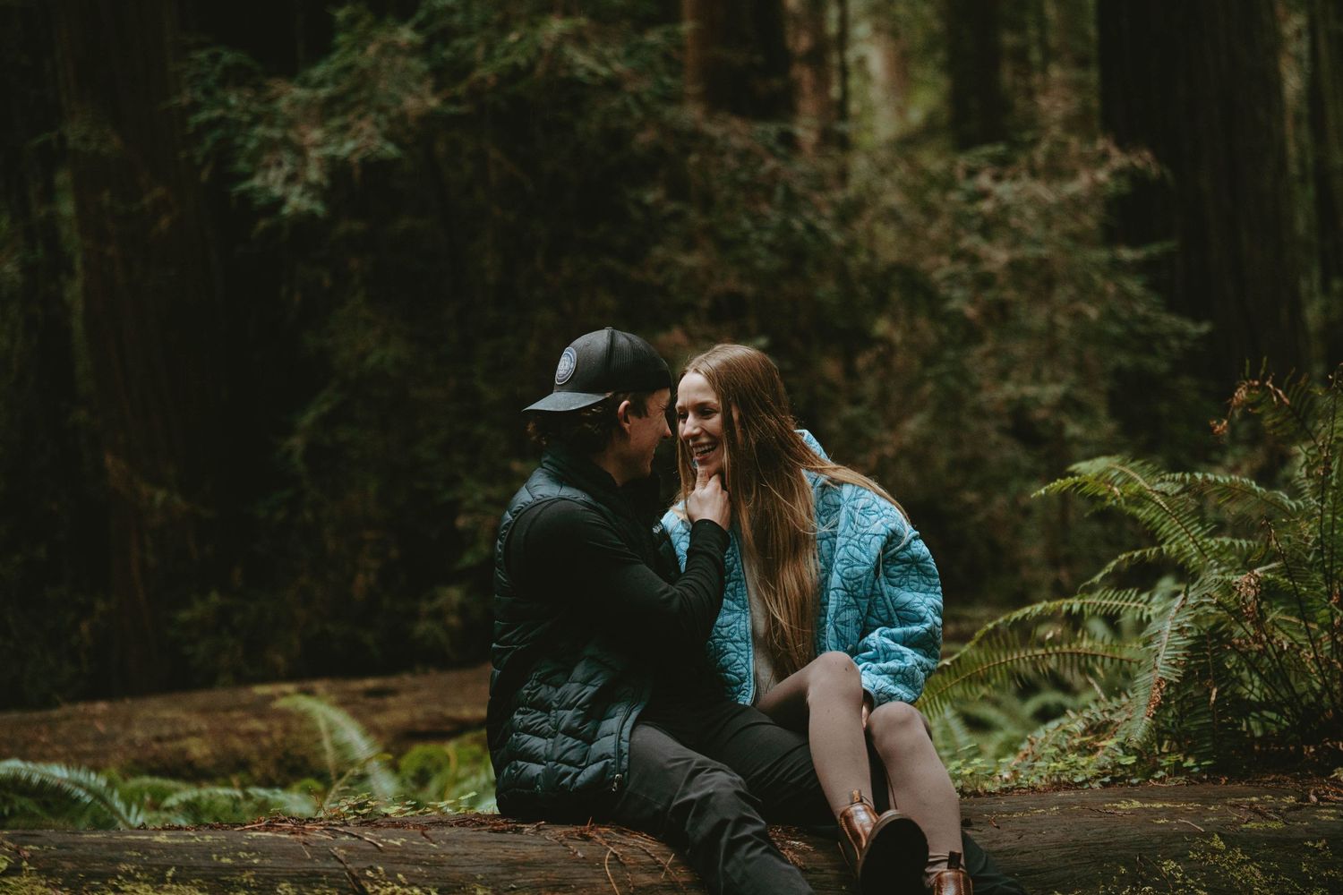 Two people share an intimate moment while sitting on a fallen log in a misty forest setting.