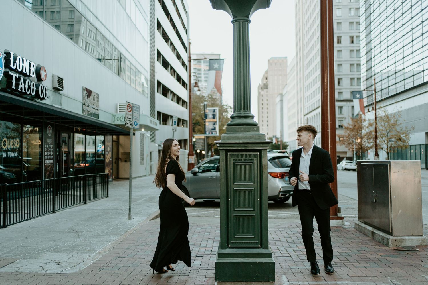 People in formal attire interact on an urban street with tall buildings and a vintage-style lamp post in downtown setting.