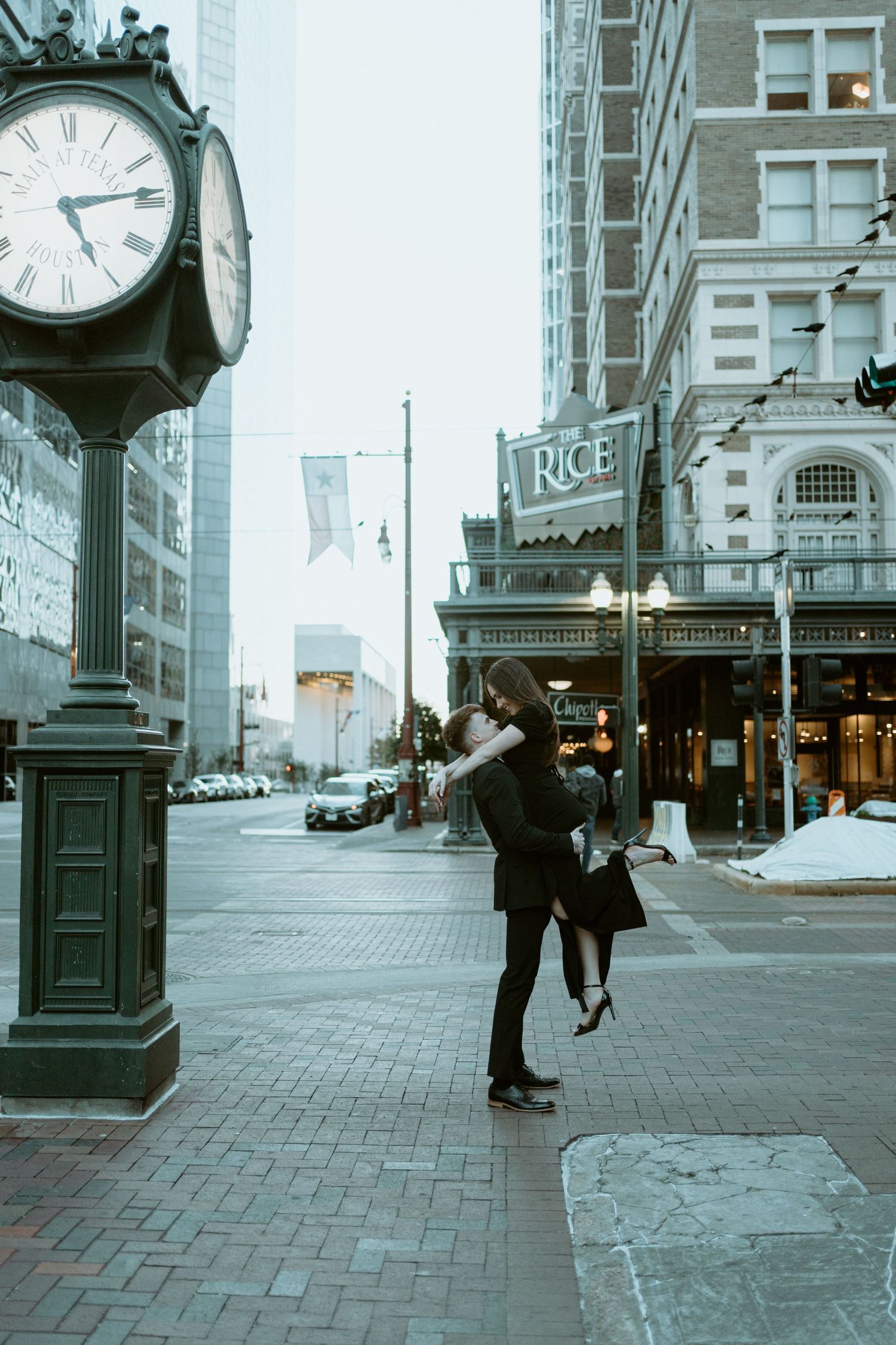 A romantic couple embraces near a vintage street clock and storefront in a moody urban setting on a winter day.