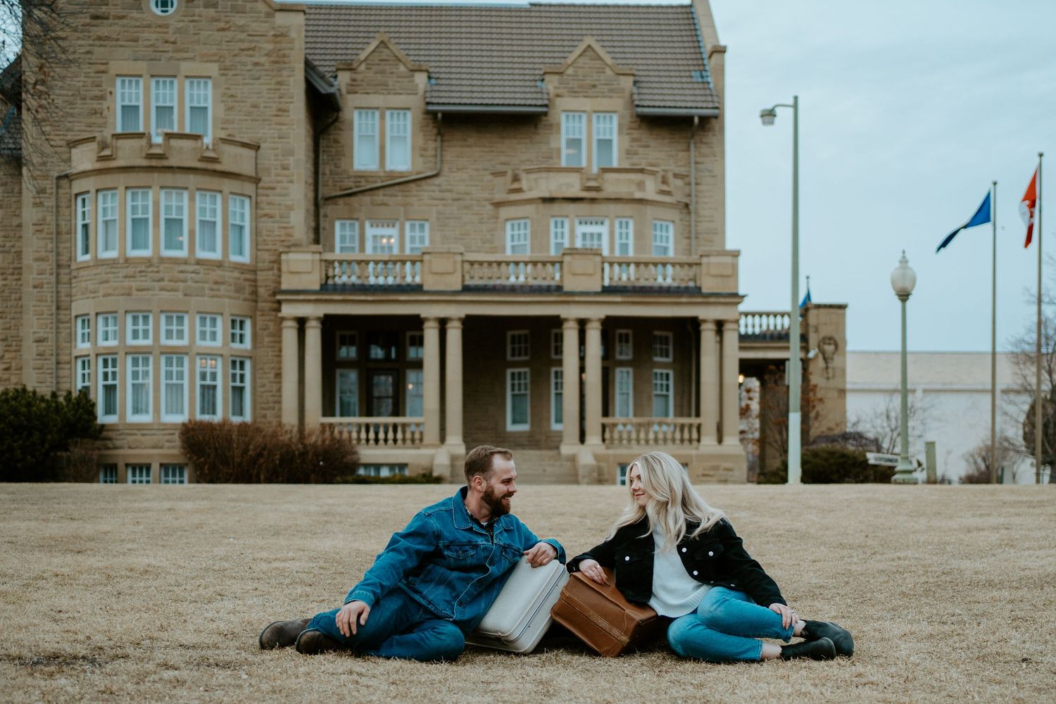 Couple relaxing together on grass in front of grand stone building with vintage luggage.