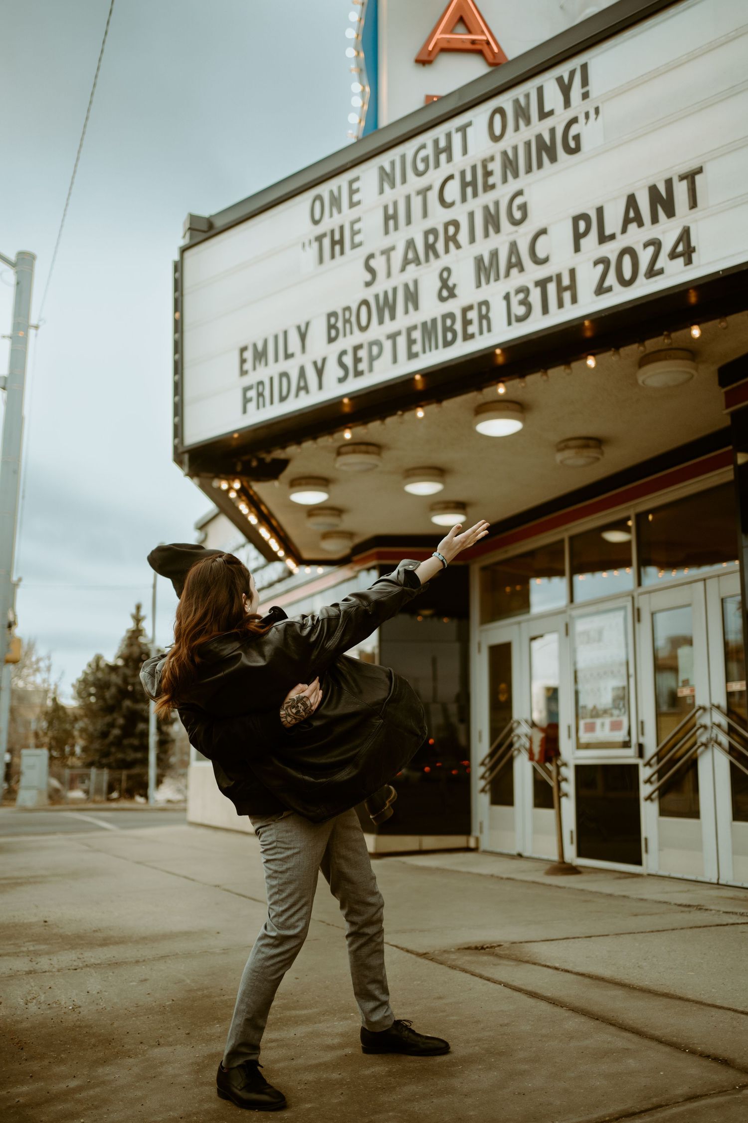A person poses dramatically in front of a vintage theatre marquee displaying upcoming shows.