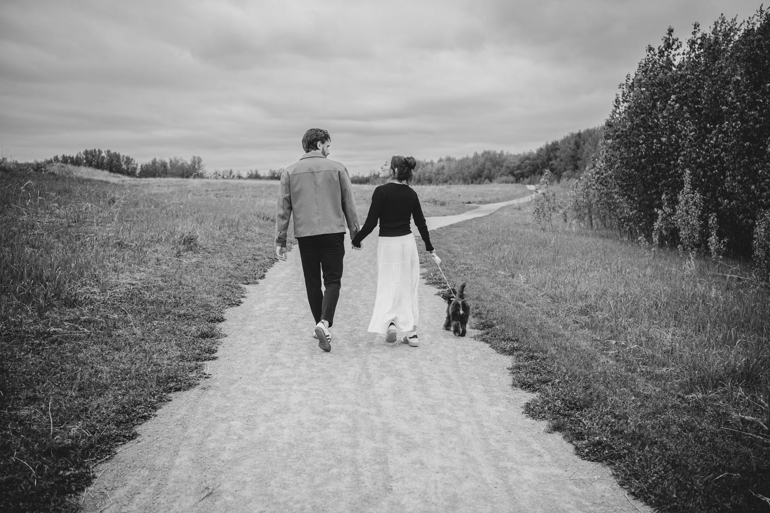 A couple walks hand in hand with their dog along a rural dirt path in black and white photography.