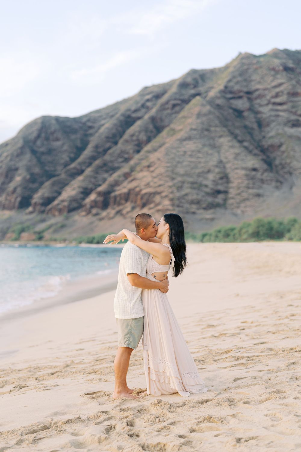 Romantic couple in white embracing on sandy beach with rugged mountain cliffs in the background.