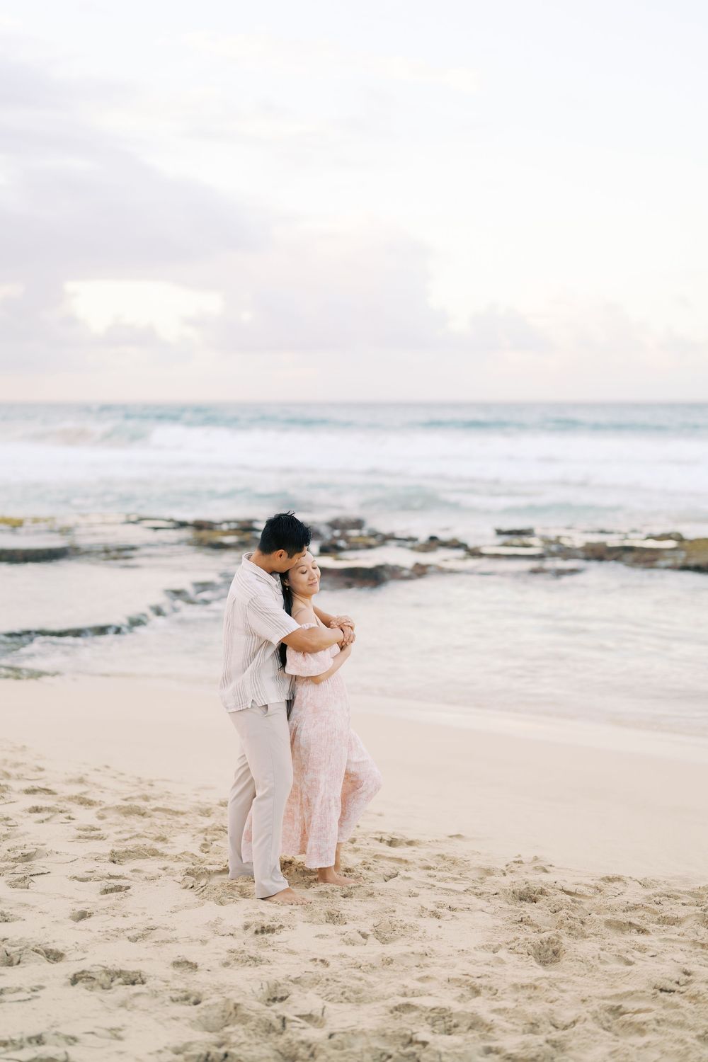 A couple in light colored outfits embrace on a misty beach with waves and rocky shoreline in the background.