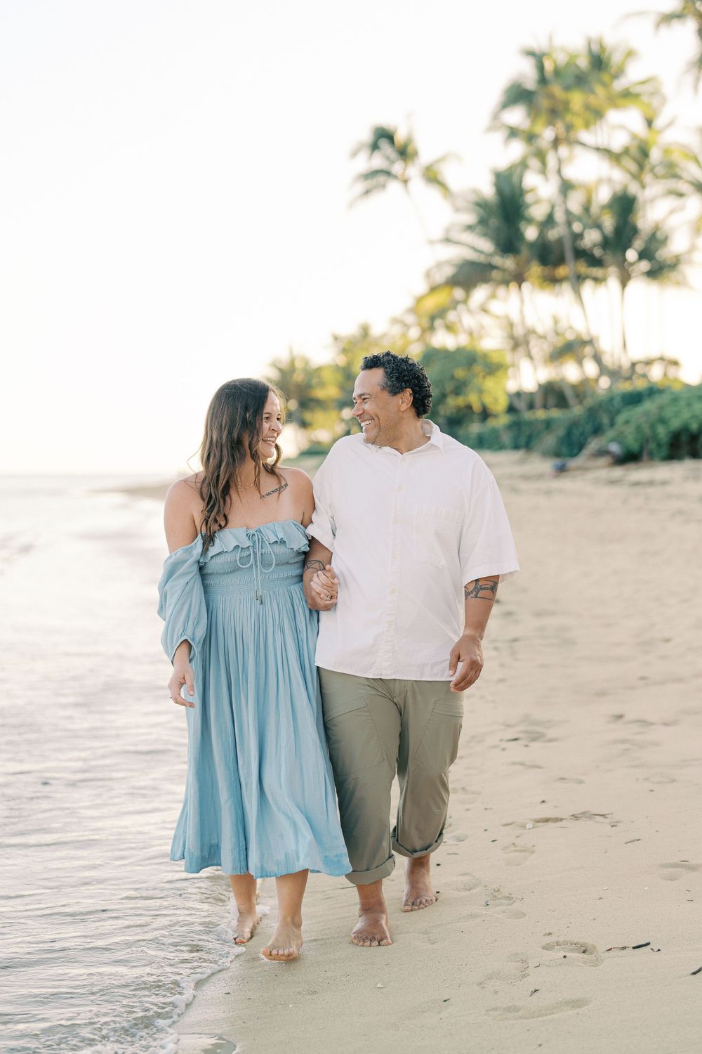 A couple in coordinated blue and white outfits share romantic moments while walking along a tropical beach at sunset.