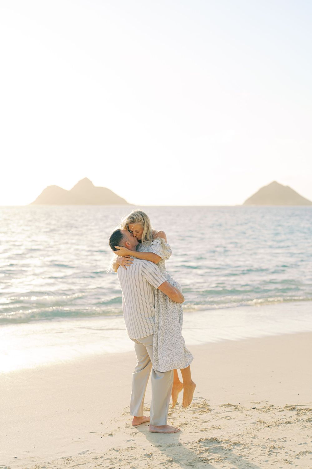 Romantic couple embracing on a sandy beach at sunset with ocean waves and mountains in the background.
