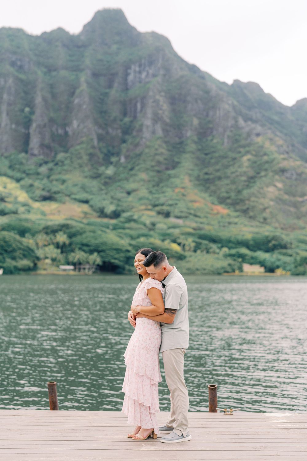 A couple embraces on a wooden dock overlooking a serene lake with dramatic mountain peaks in Hawaii.