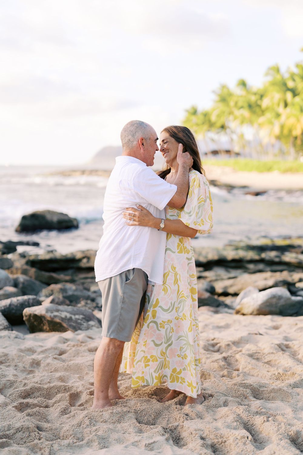 A couple shares a romantic moment on a sandy beach with black rocks and palm trees in the background at sunset.
