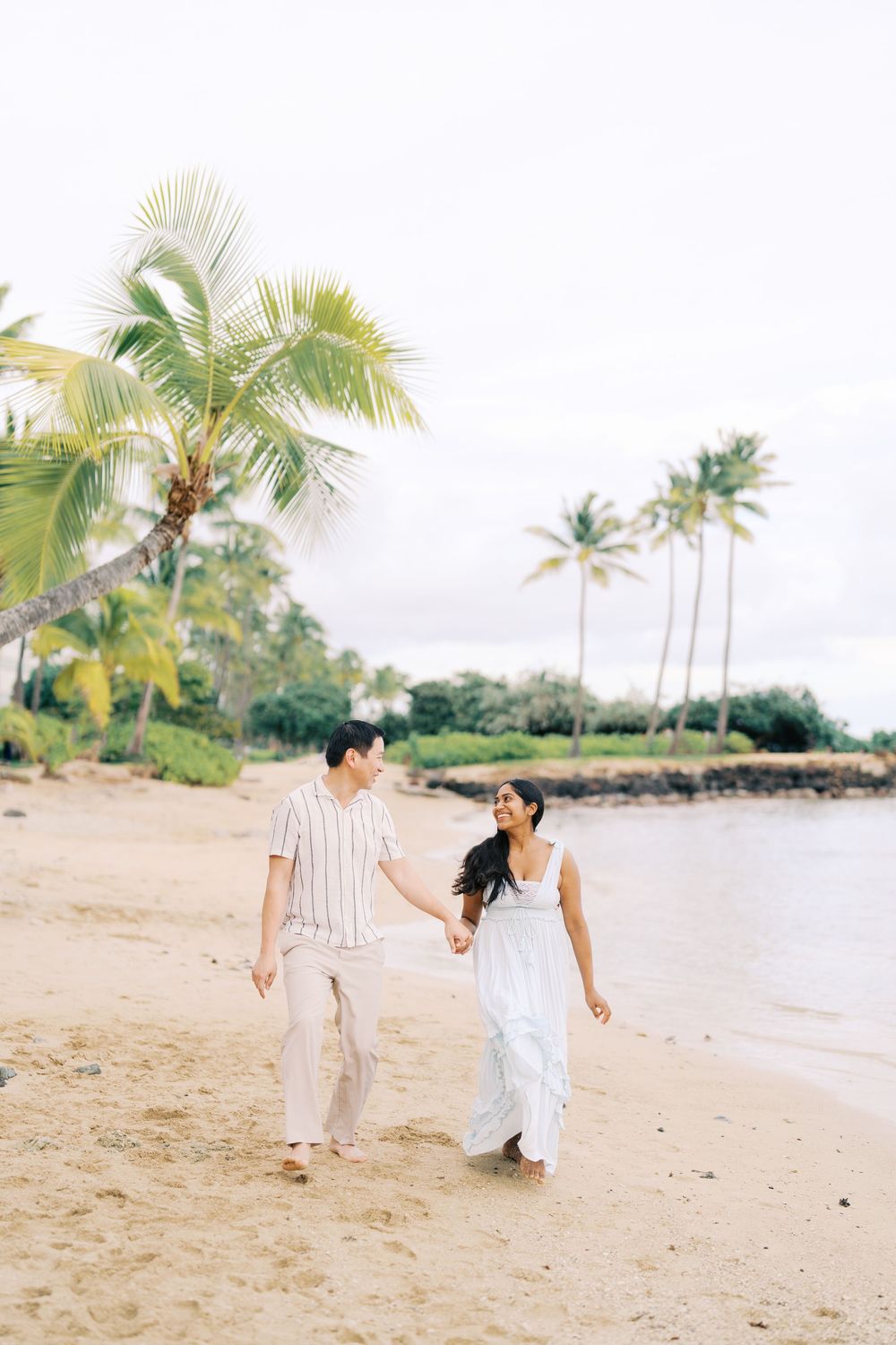 A couple walks hand in hand along a tropical beach with palm trees and ocean views in Hawaii.