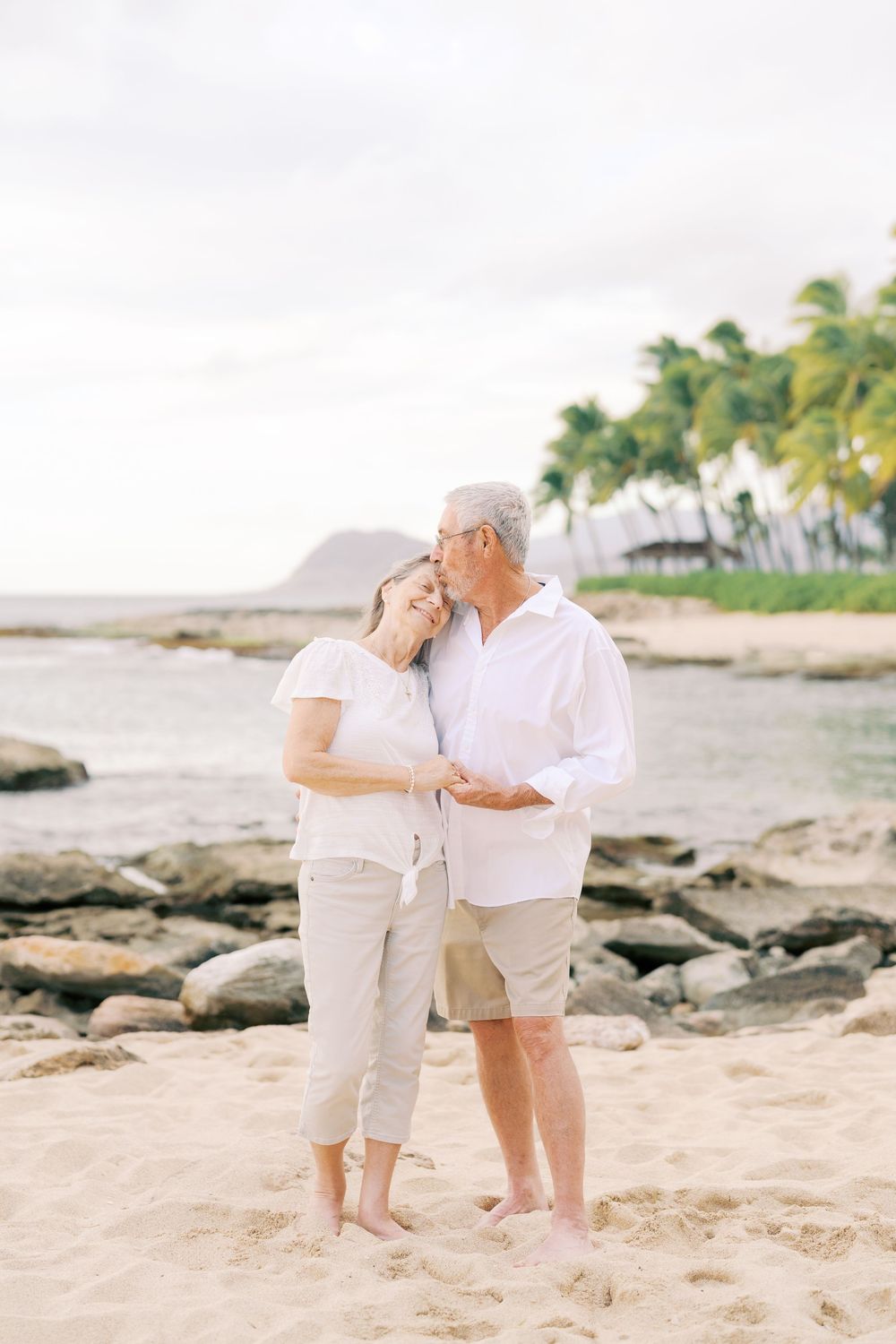 A couple in white clothing shares a romantic moment on a tropical beach with palm trees and rocks in the background.