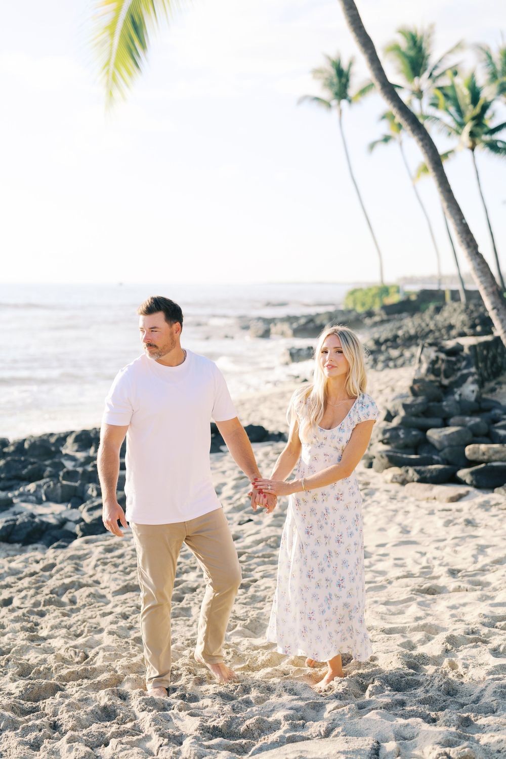 Couple walking hand in hand on a tropical beach with palm trees and black lava rocks in Hawaii.
