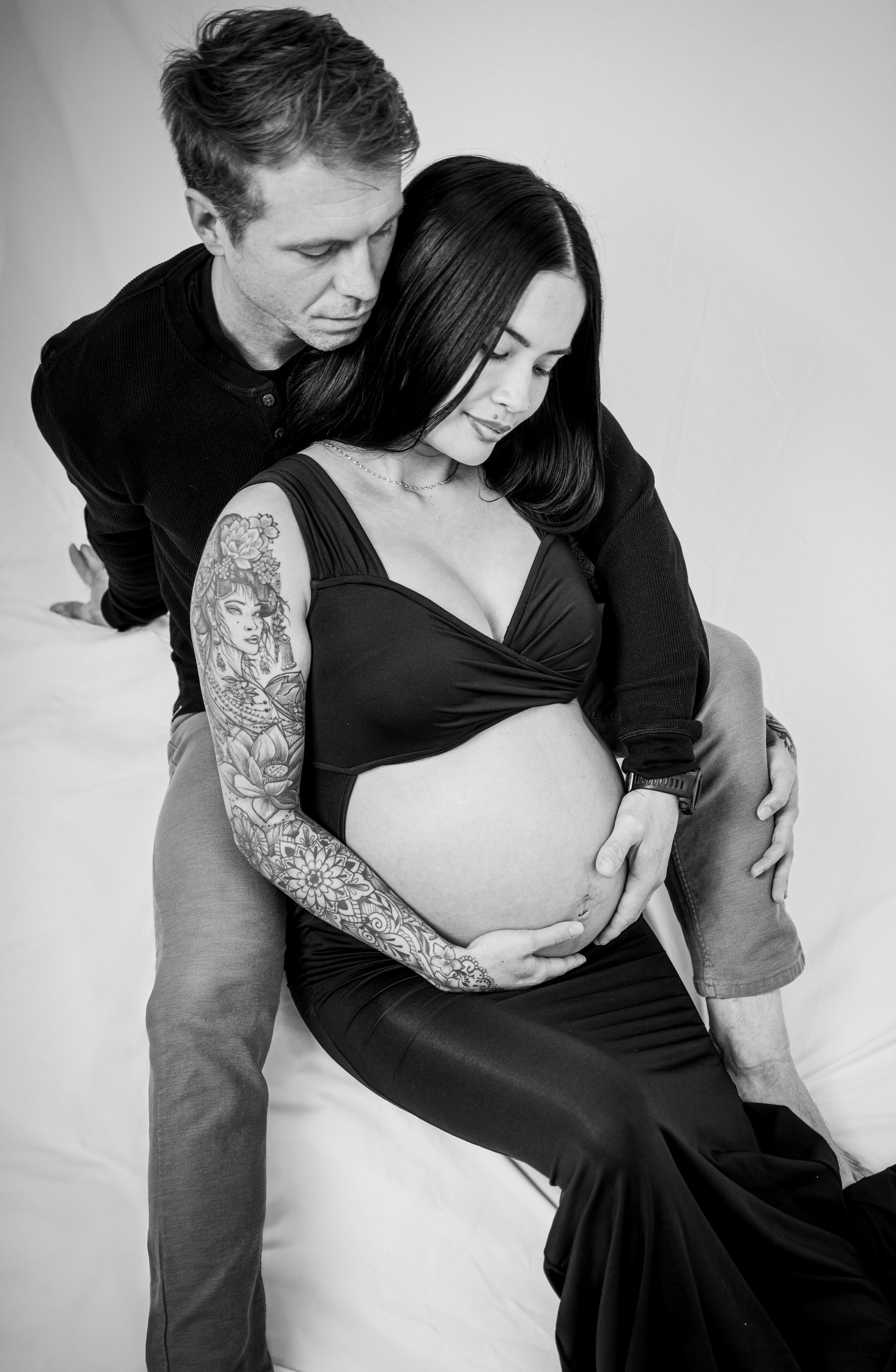 Artistic black and white maternity photo showing a couple embracing, with the woman wearing a long dress and displaying sleeve tattoos.