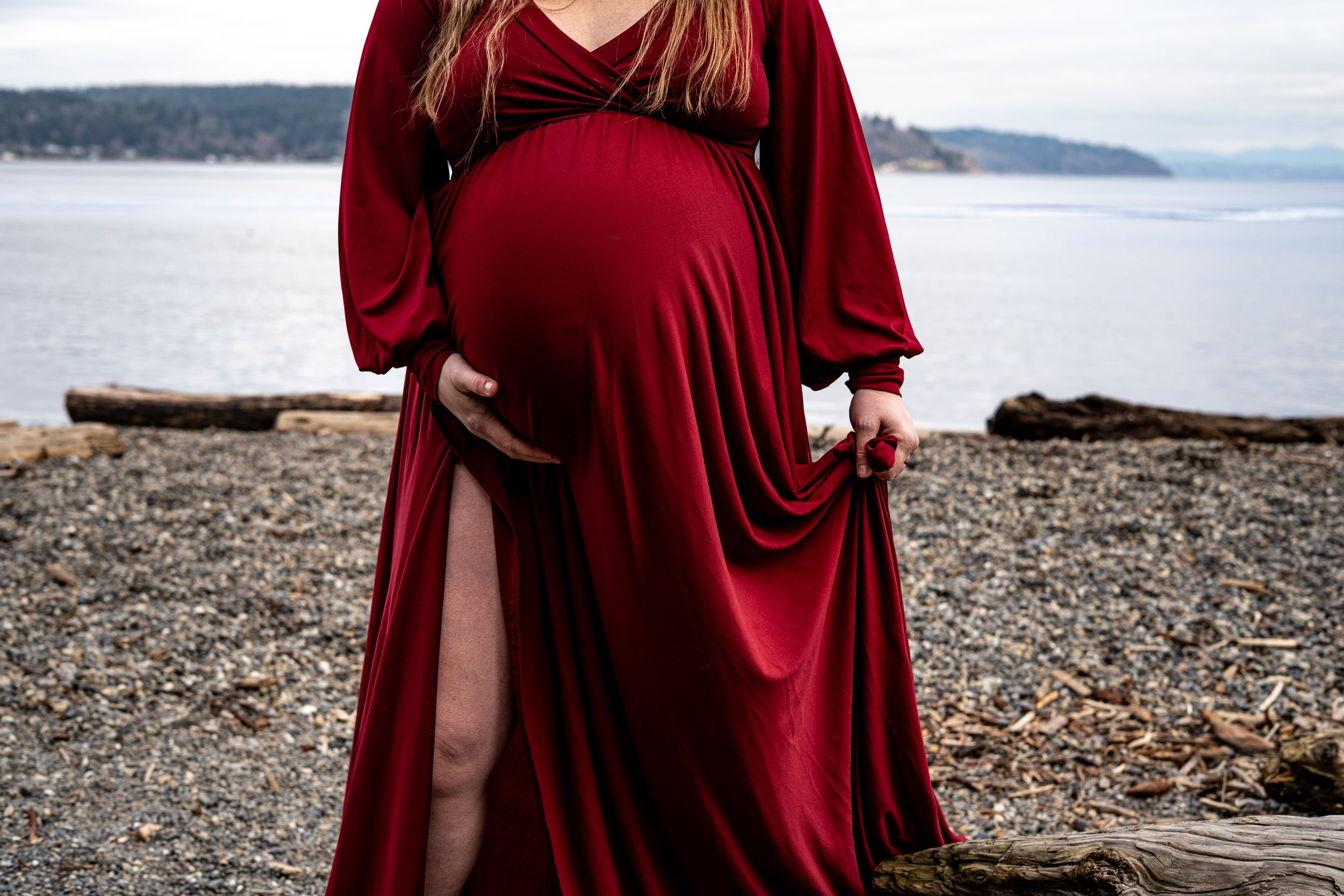 A person in a flowing burgundy dress walks along a rocky beach shoreline with mountains in the distance.