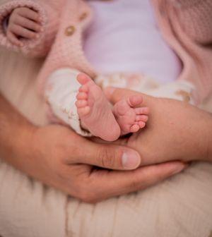 Adult hands gently cradling tiny newborn baby feet, dressed in a soft pink outfit.