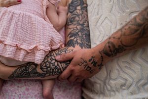 Tattooed hand gently touching small feet wearing pink ruffled clothing on a patterned gray background.