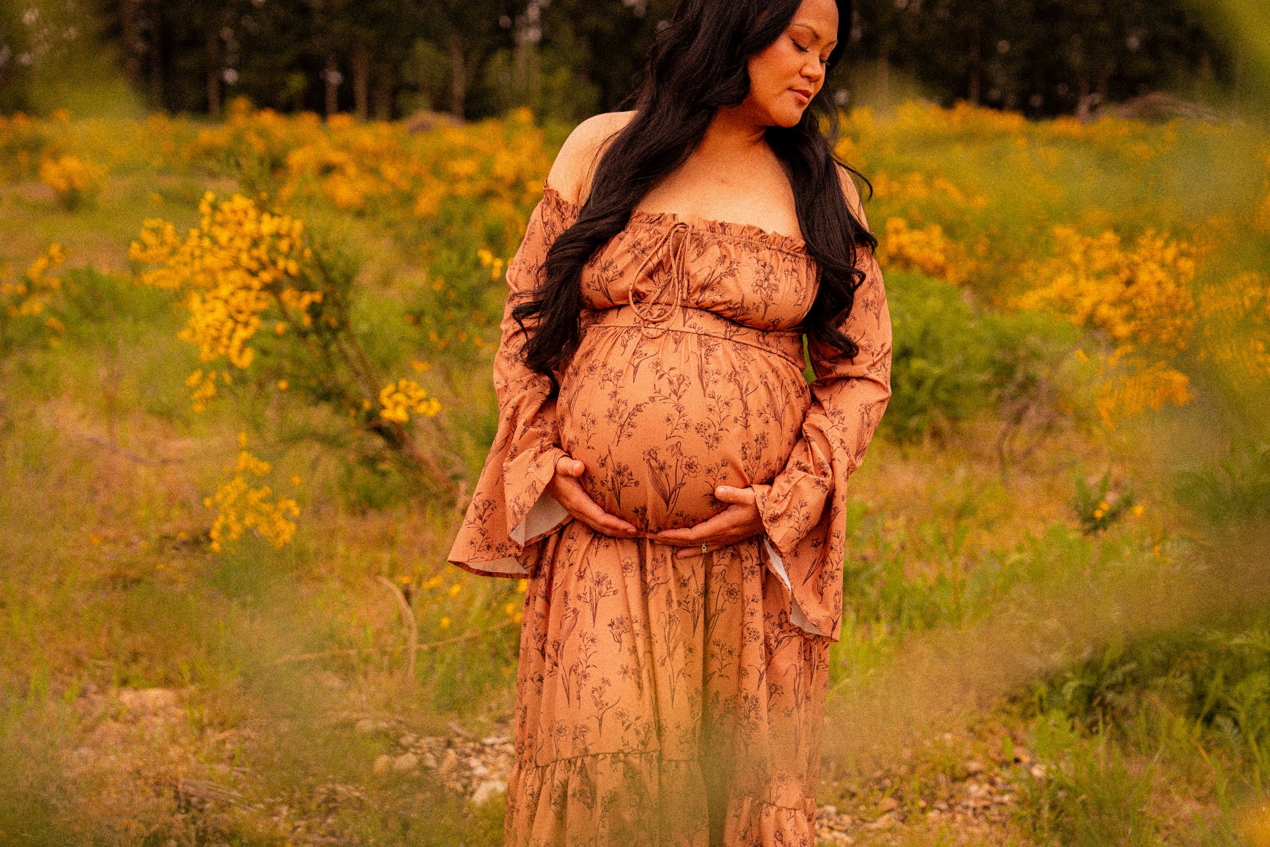 Pregnant woman in floral maxi dress holding baby bump in golden wildflower field during maternity photo session.