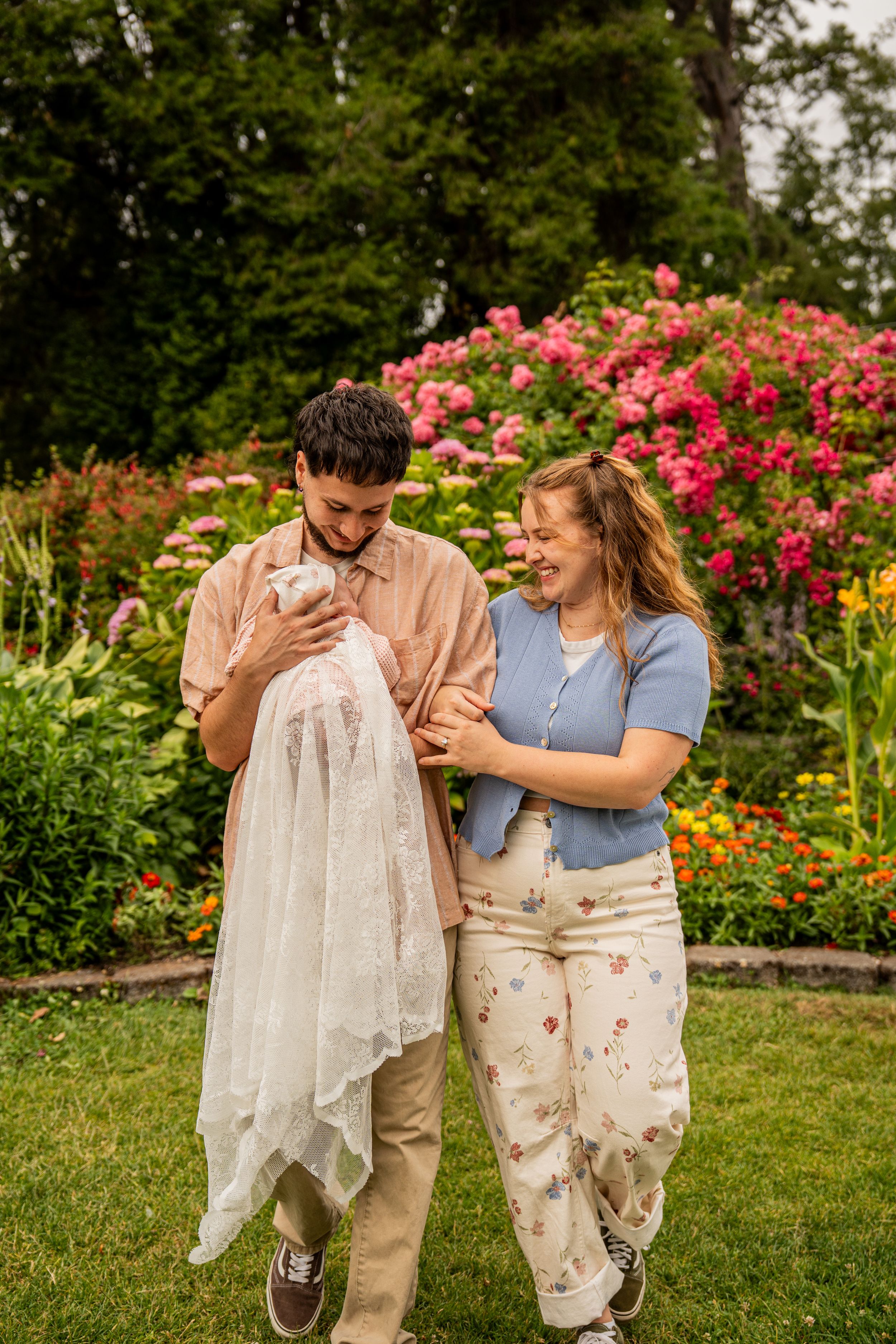 A couple smiles at their newborn baby in a lush garden with colorful blooming flowers.