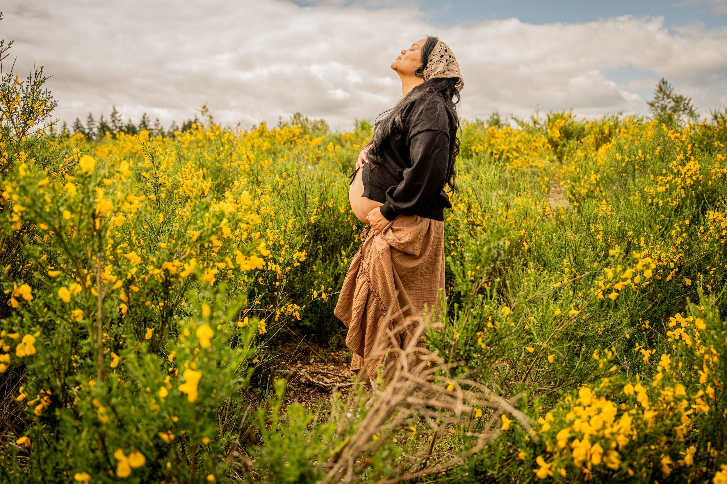 Pregnant woman in vintage attire standing in a vibrant yellow wildflower field under a cloudy sky.