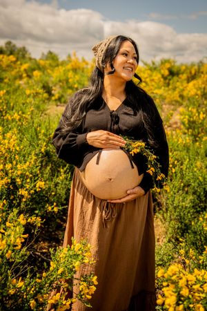 Smiling pregnant woman in black top and brown skirt holding belly in yellow flower field outdoors.