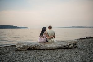 A romantic couple sits on a rocky beach at sunset overlooking a serene ocean view.