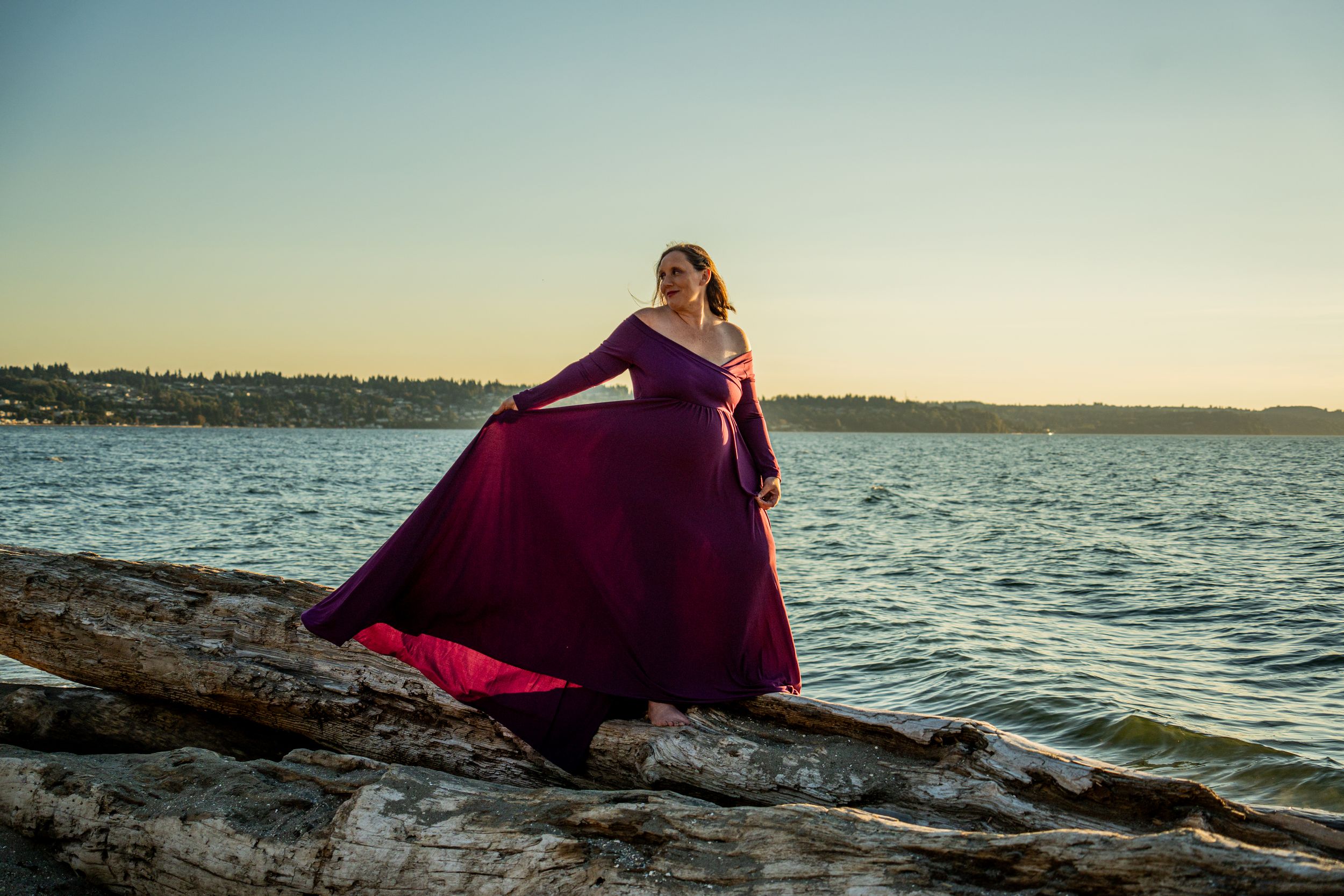 A flowing burgundy gown catches the wind as someone poses on rocky shoreline at sunset.
